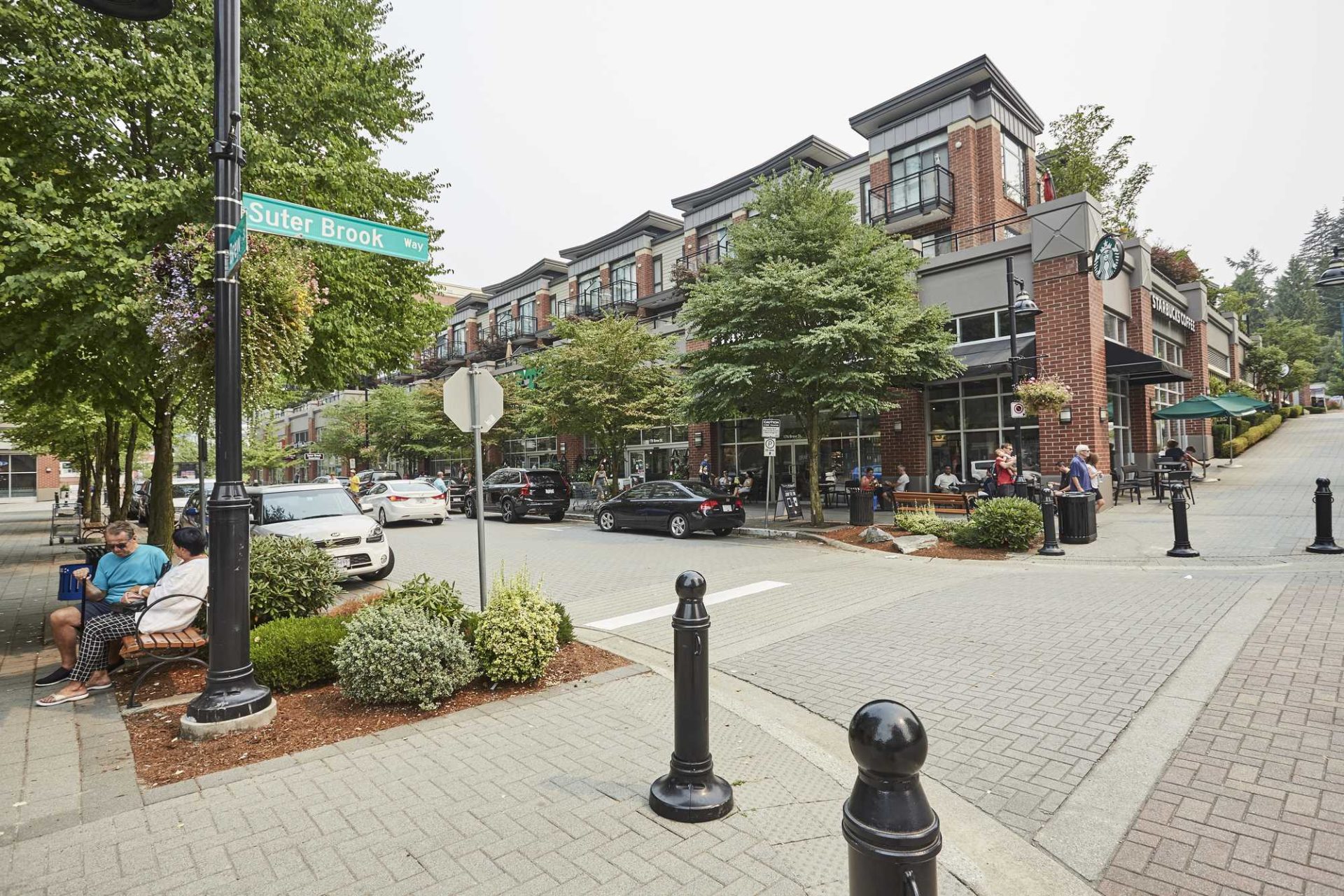  Street view of Suter Brook Village in Port Moody featuring pedestrians, parked cars, trees, and Starbucks on a sunny day.